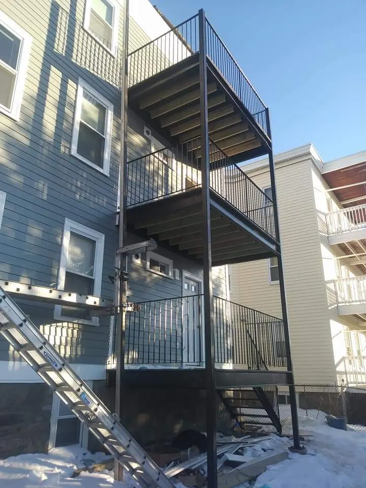 Three stacked steel balconies with black railings on the rear of a gray clapboard building