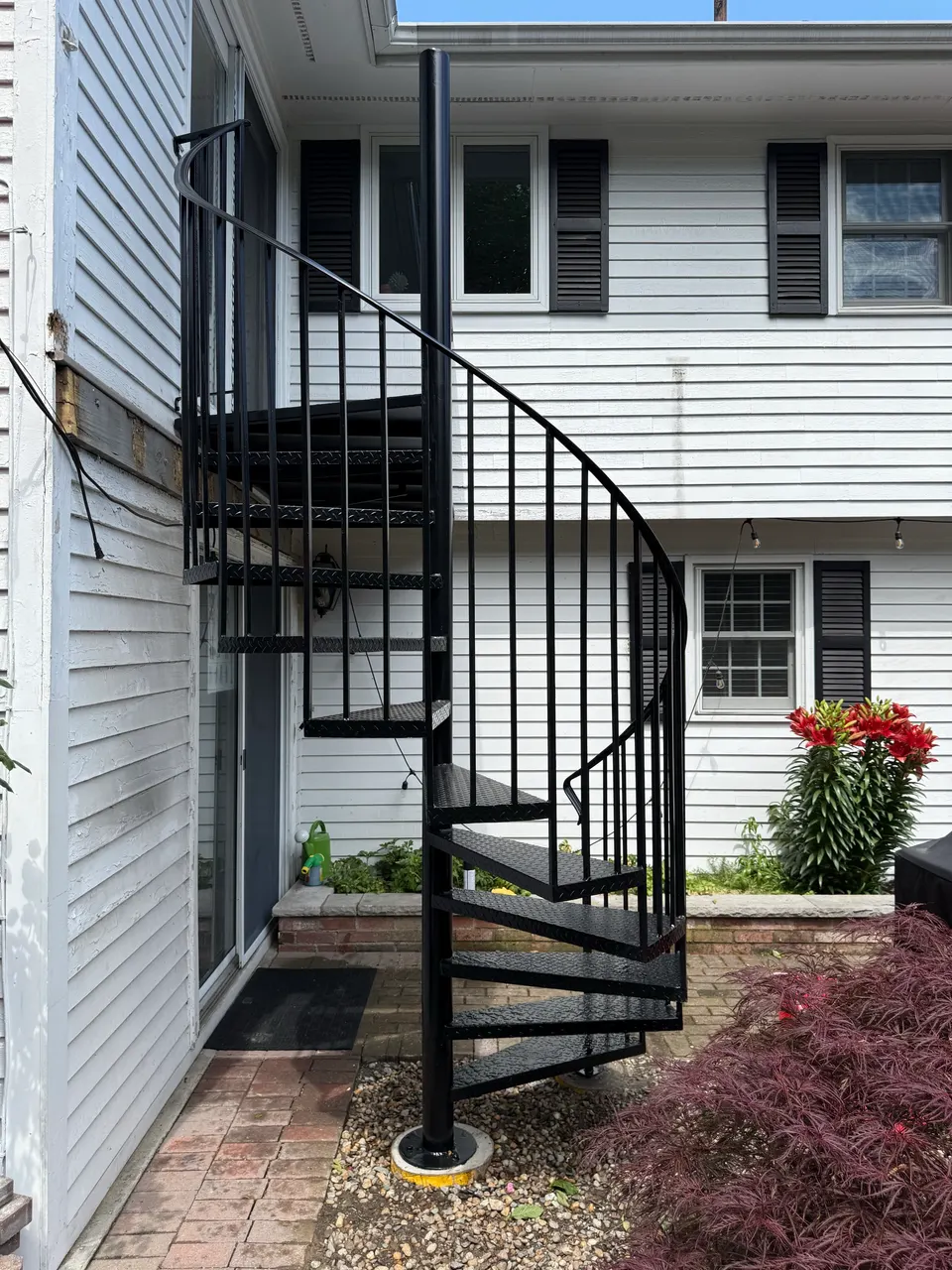 Black spiral stair with vertical balusters rising to an upper door beside a patio